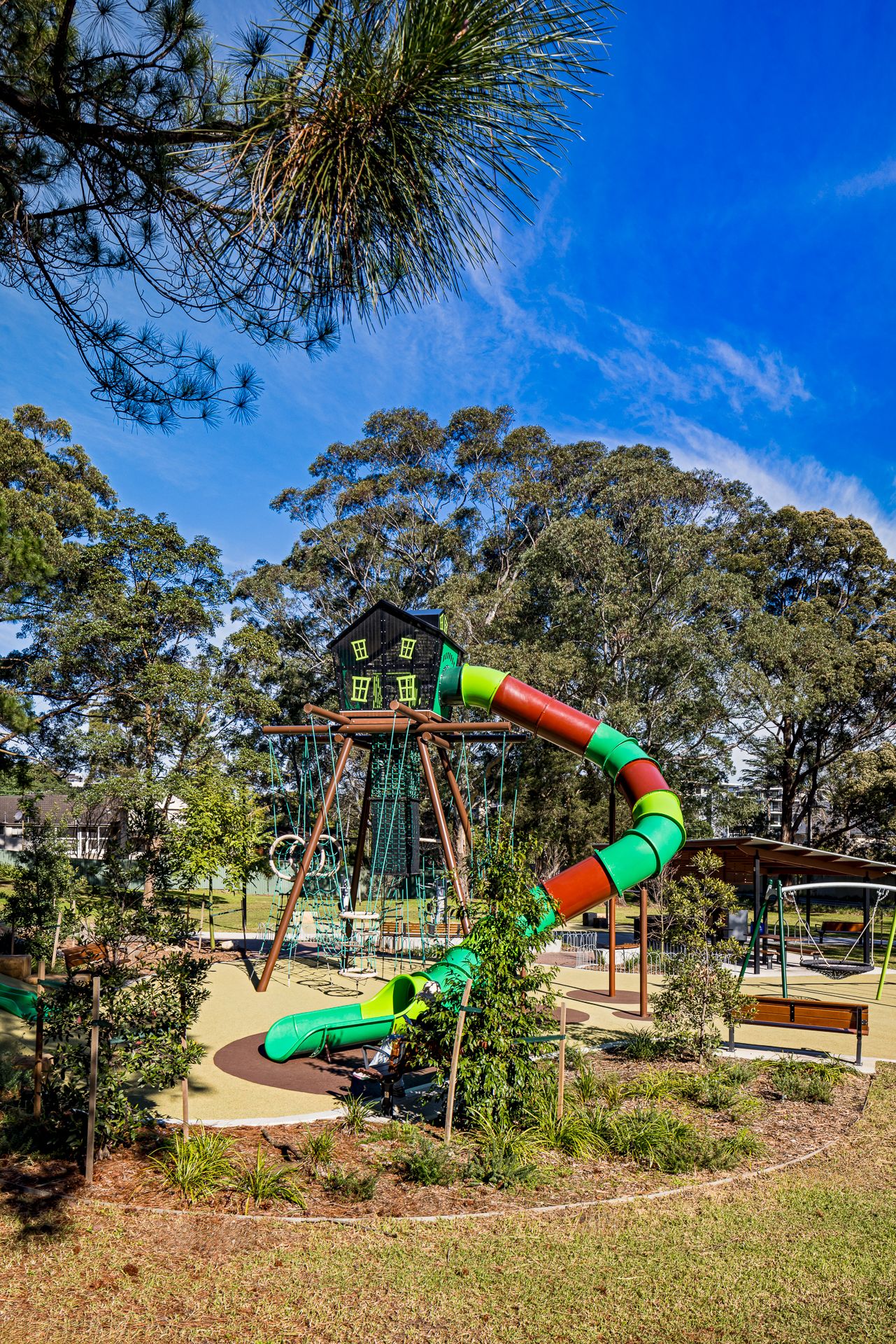 Colorful green, red and orange tube slide attached to a wooden treehouse play tower surrounded by eucalyptus trees under a bright blue sky.