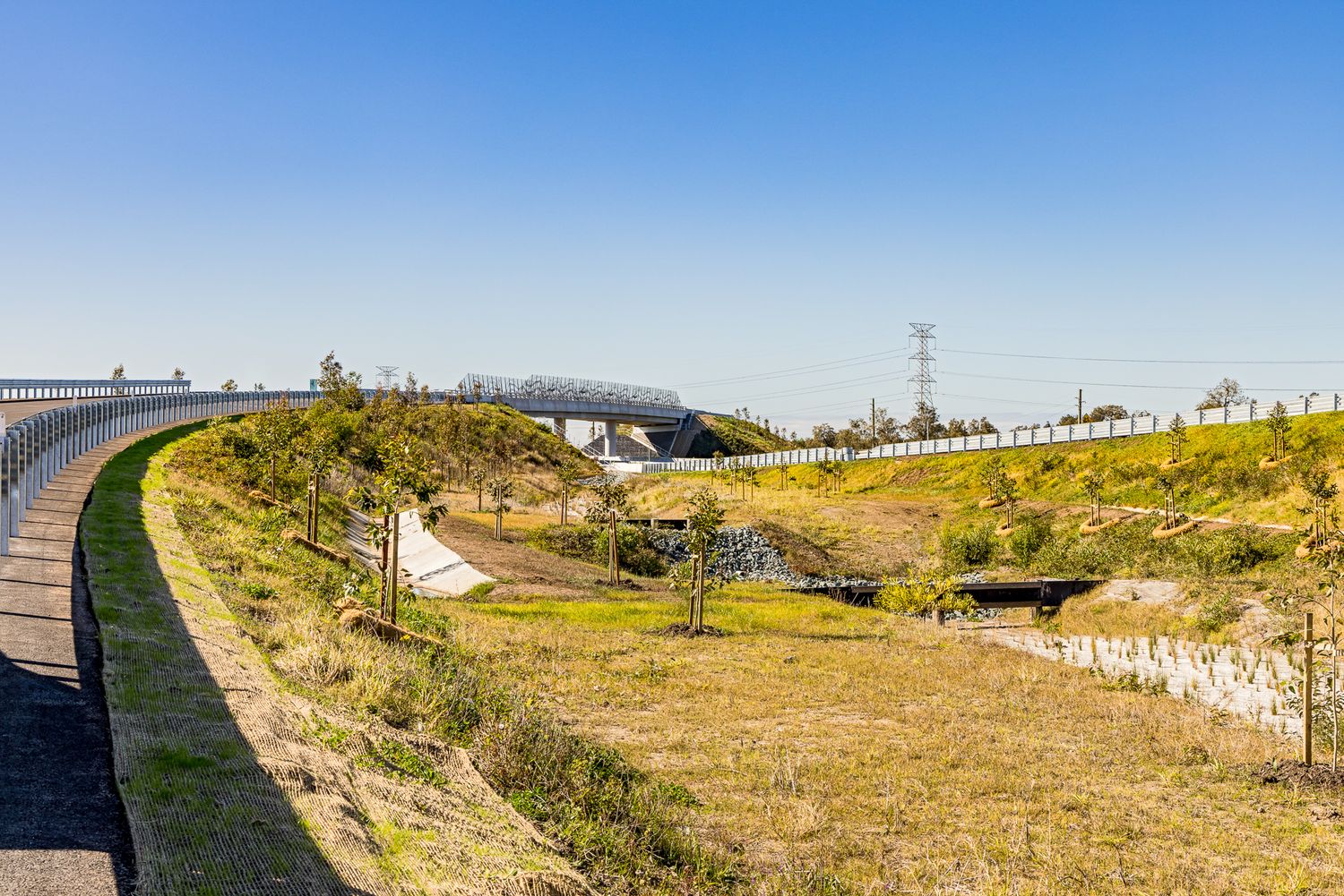 Curved new motorway lane with concrete barrier beside grassy embankments and an overpass under a clear blue sky.