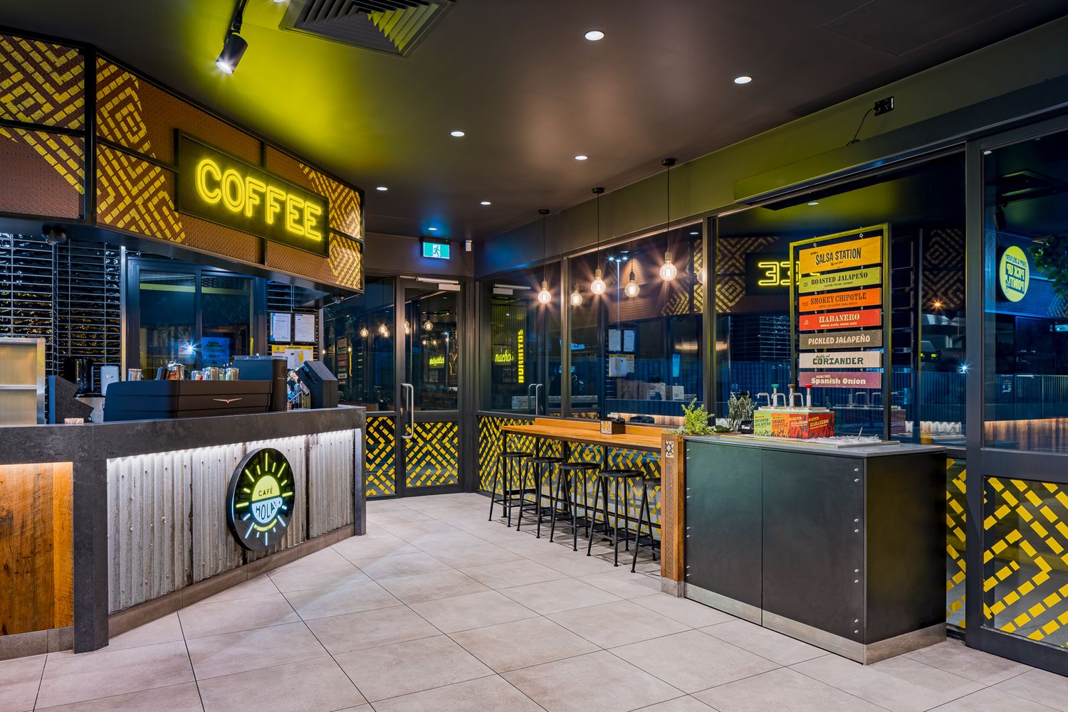 Restaurant interior with an illuminated coffee sign, timber bar stools, geometric yellow patterned walls and a service counter.