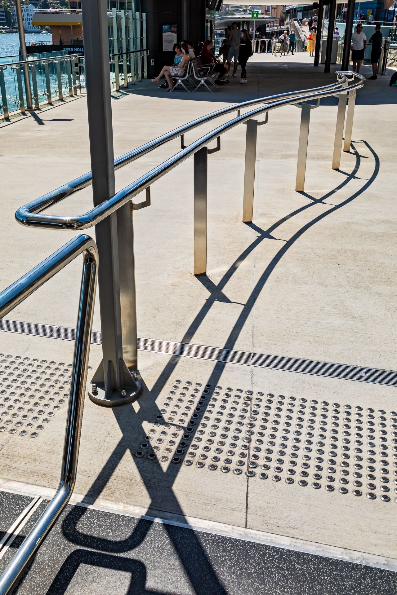 Curved stainless steel handrails cast long shadows across a ferry wharf walkway with tactile paving, as passengers sit in the background.