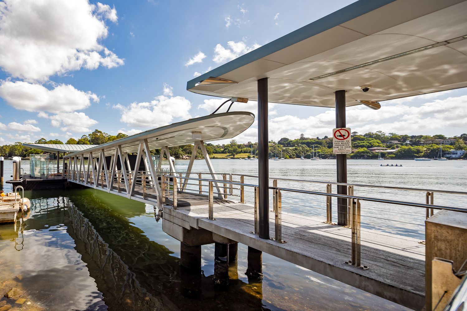 Timber ferry wharf with canopy roof and steel gangway extending over calm harbour water, with tree-lined shores under a blue sky.