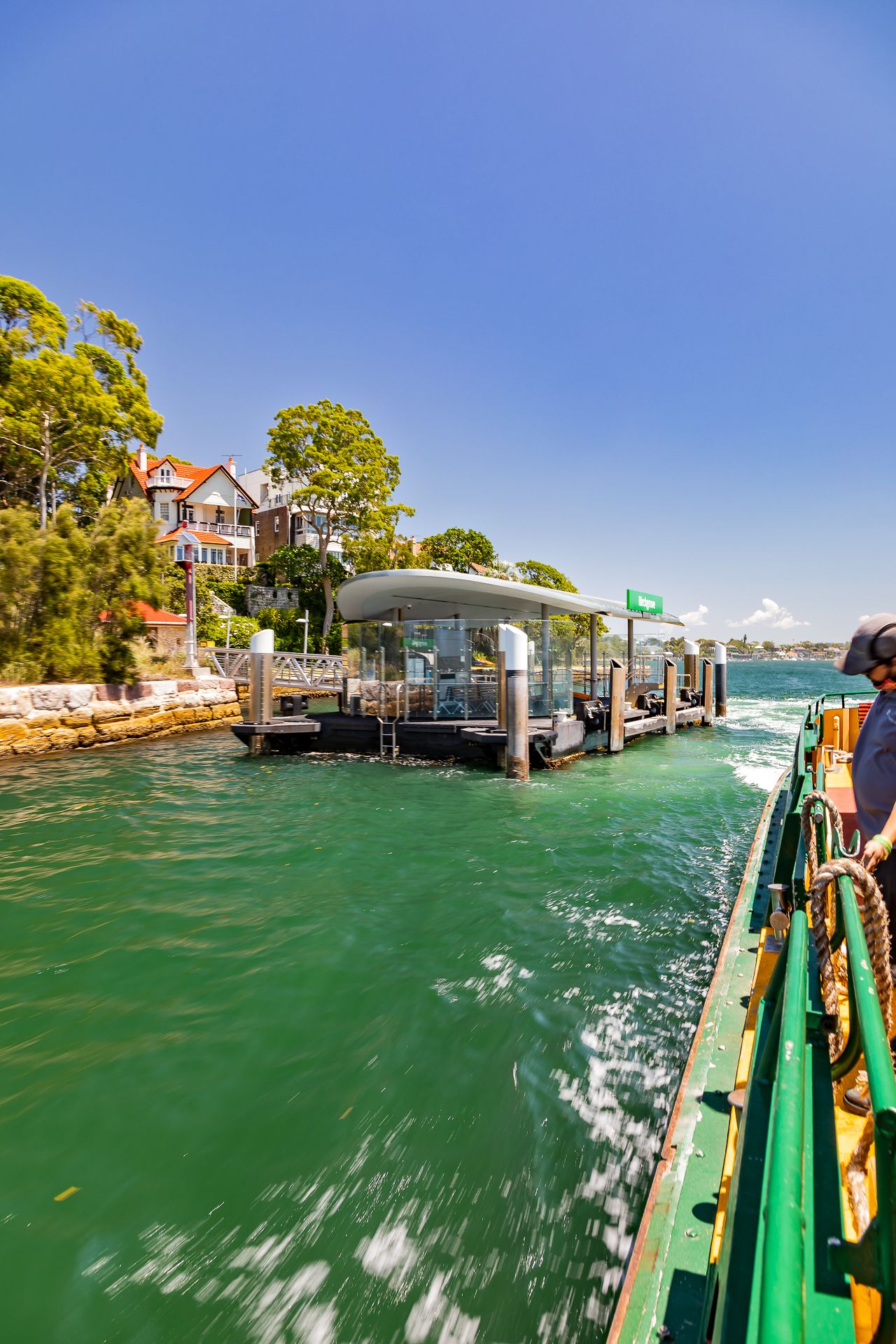 Ferry deck view of a modern covered wharf beside a waterfront house and lush trees on a sunny day with green harbour water.