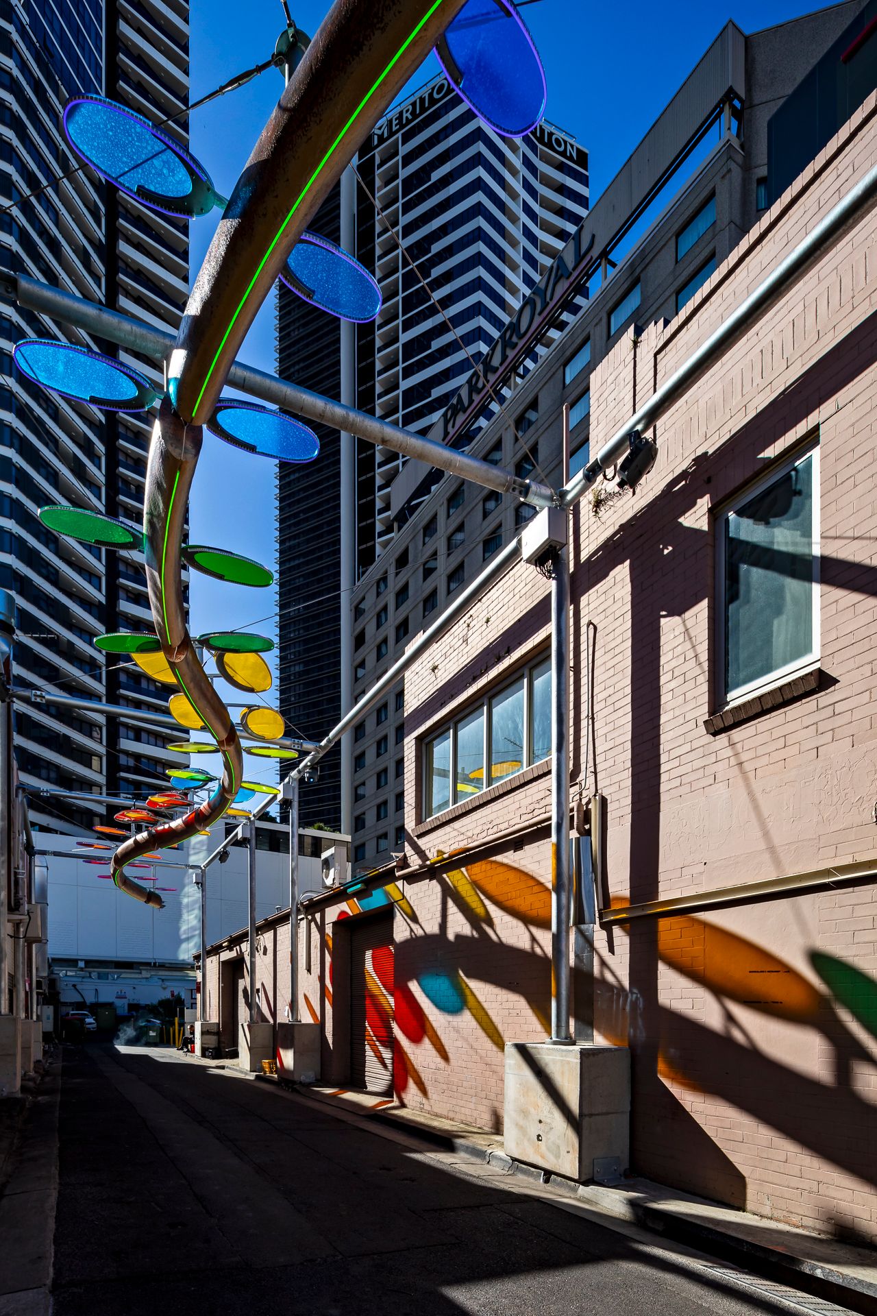 Low-angle view of a suspended sculpture with vivid blue, green and orange discs above a laneway, casting colorful shadows on brick.