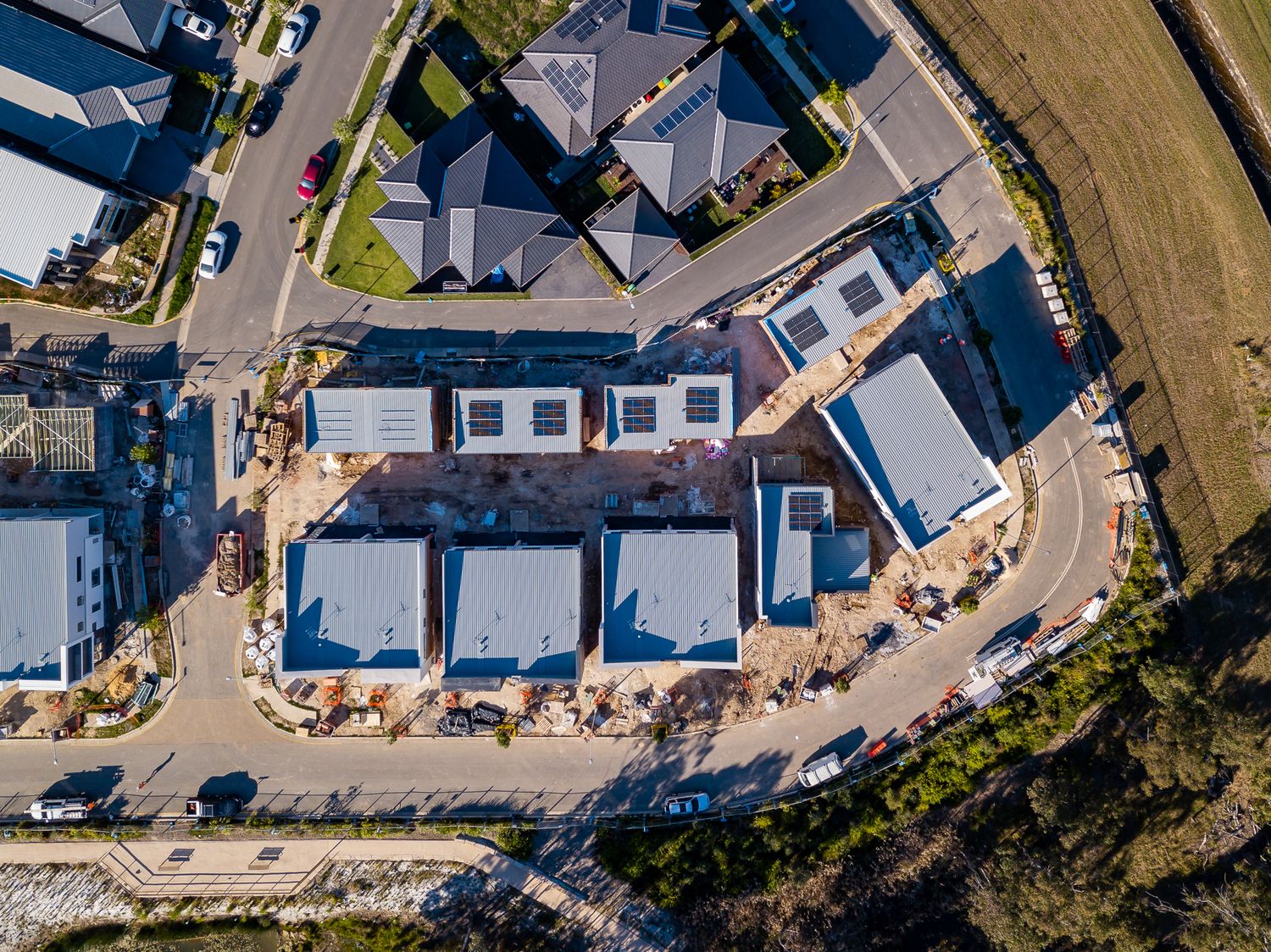 Overhead drone view of townhouse developments with new blue roofs on a curved street beside grassland in a growing residential area.