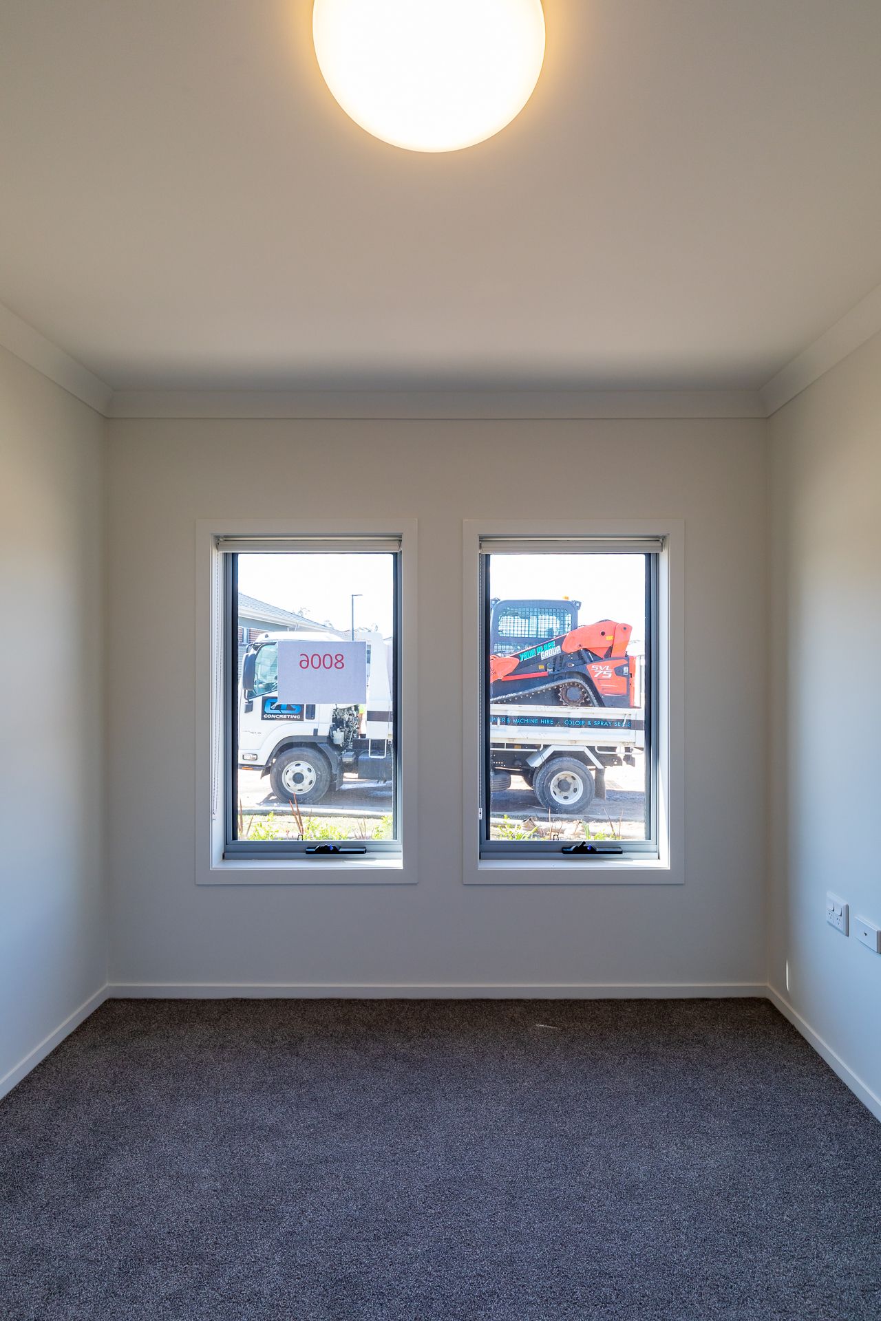 Small carpeted bedroom with two windows overlooking a truck and orange machinery on the street outside.