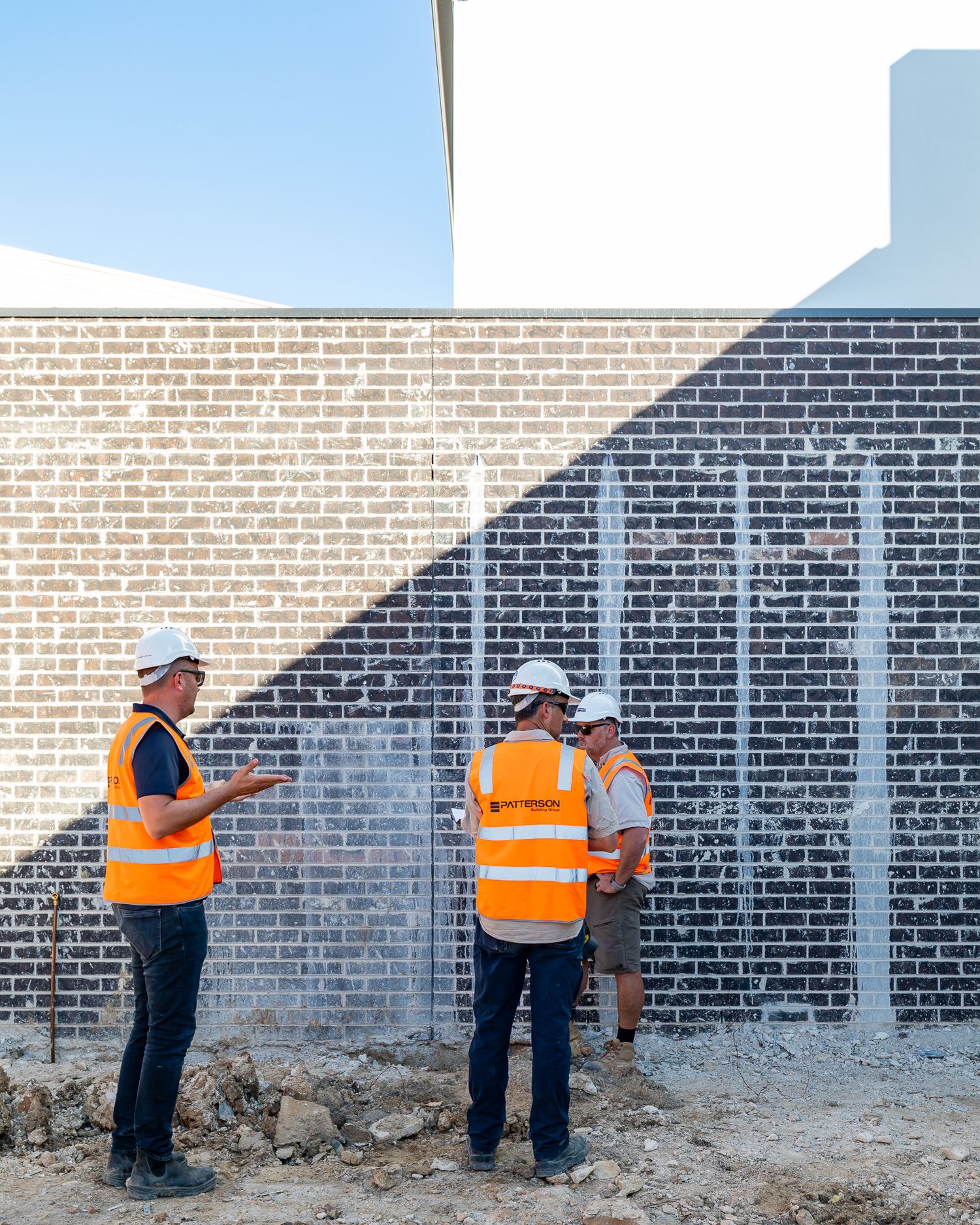 Three construction workers in hi-vis vests and white hard hats discuss progress in front of a freshly built brick wall.