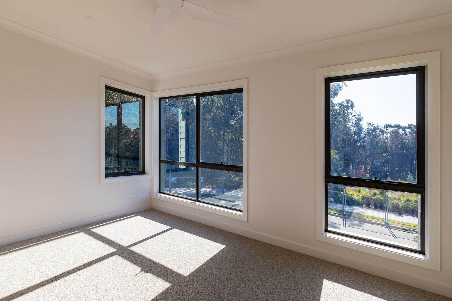 Empty bedroom in a new home with large black-framed windows, carpeted floor and sunlight casting shadows over white walls.