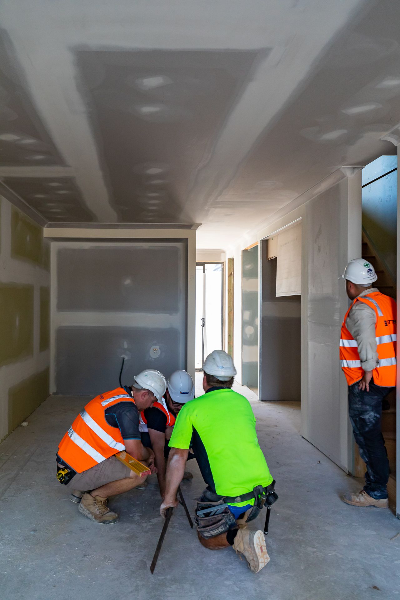 Builders in hard hats and orange safety vests gather in a hallway with fitted plasterboard walls during a new home construction inspection.