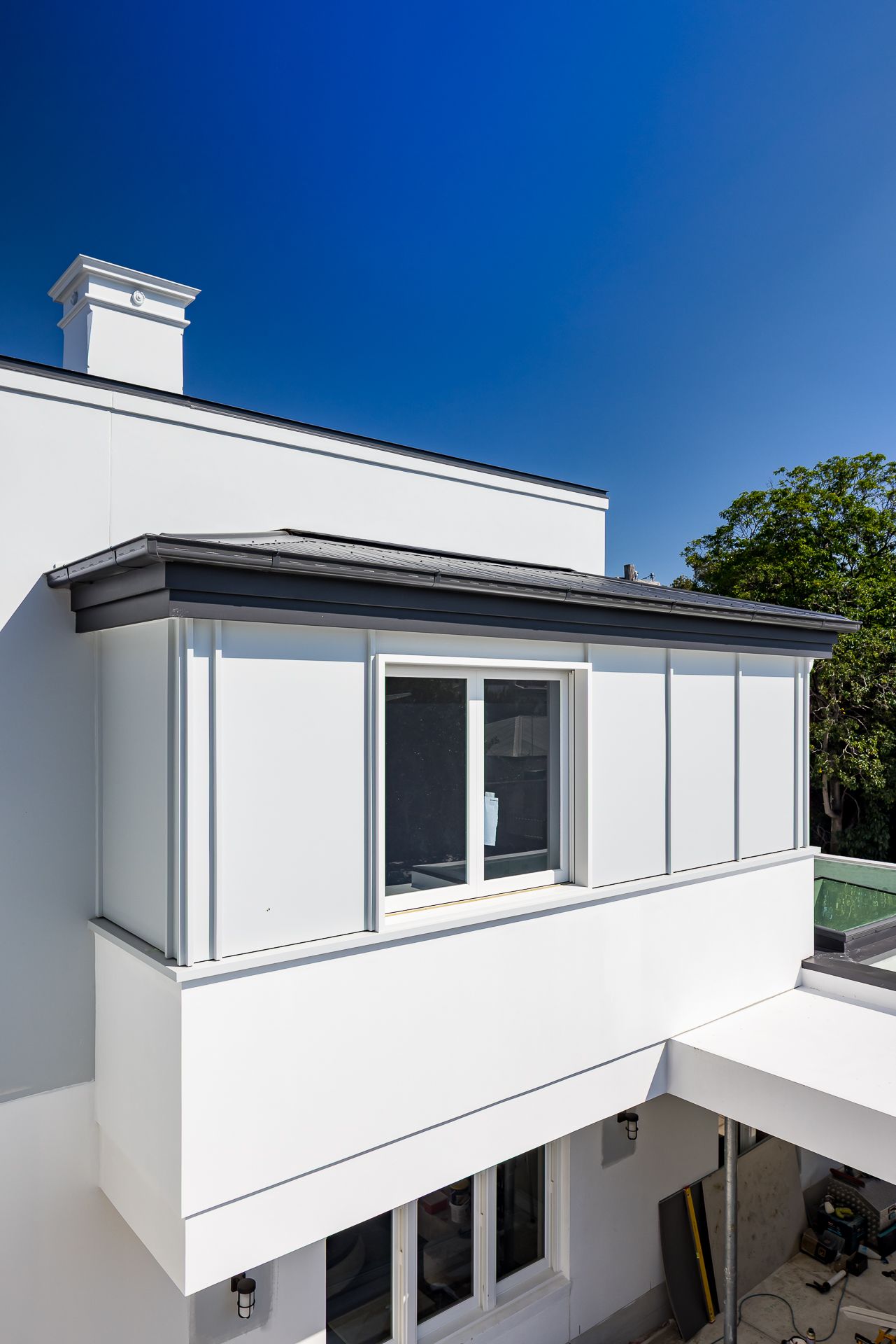 Contemporary white and grey house exterior with a cantilevered upper level, framed window, chimney and clear blue sky.