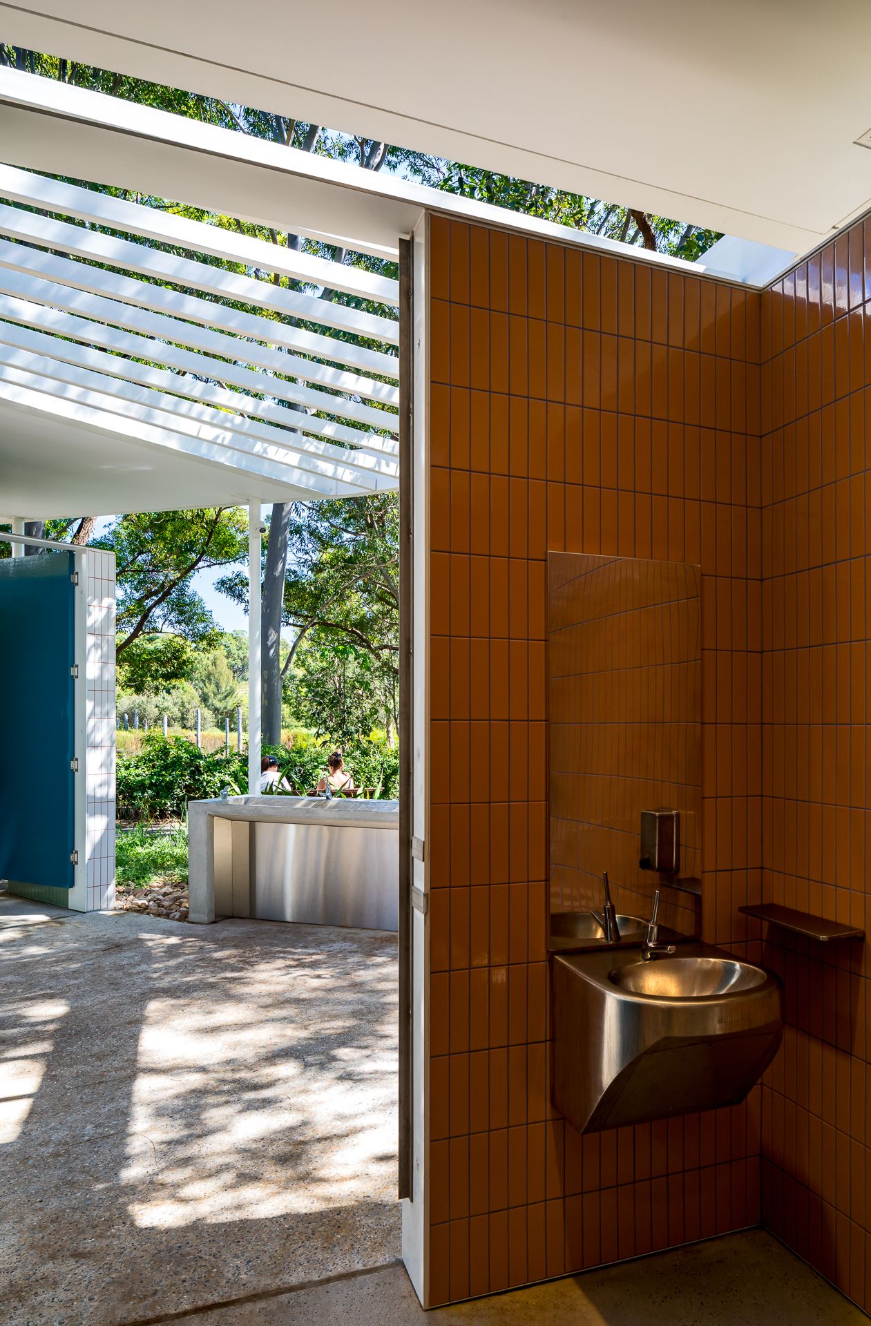 Open-air restroom with orange ceramic tiles, a stainless steel sink and a blue door overlooking a sunny garden.