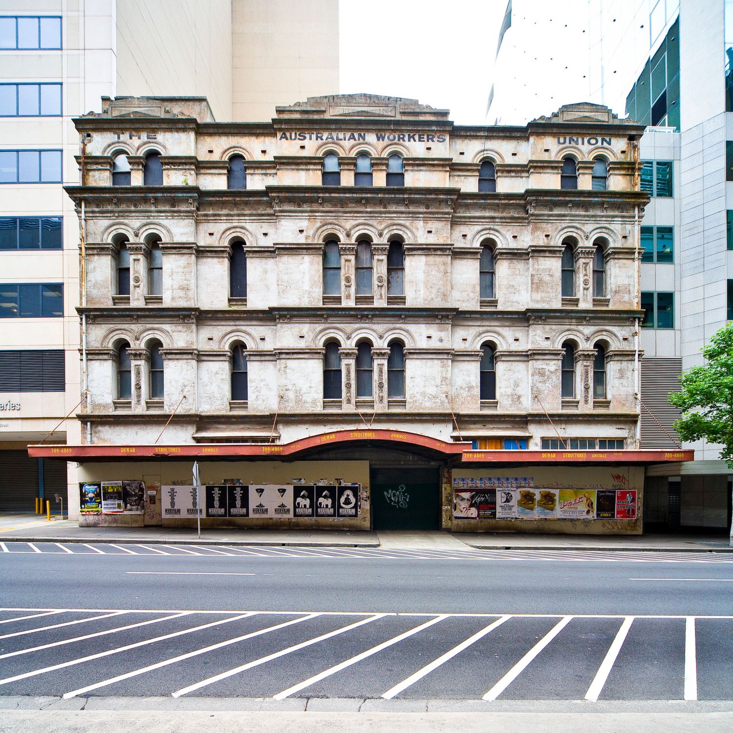 Ornate Victorian-era building facade with arched windows and a red awning, standing between modern glass towers in the city.