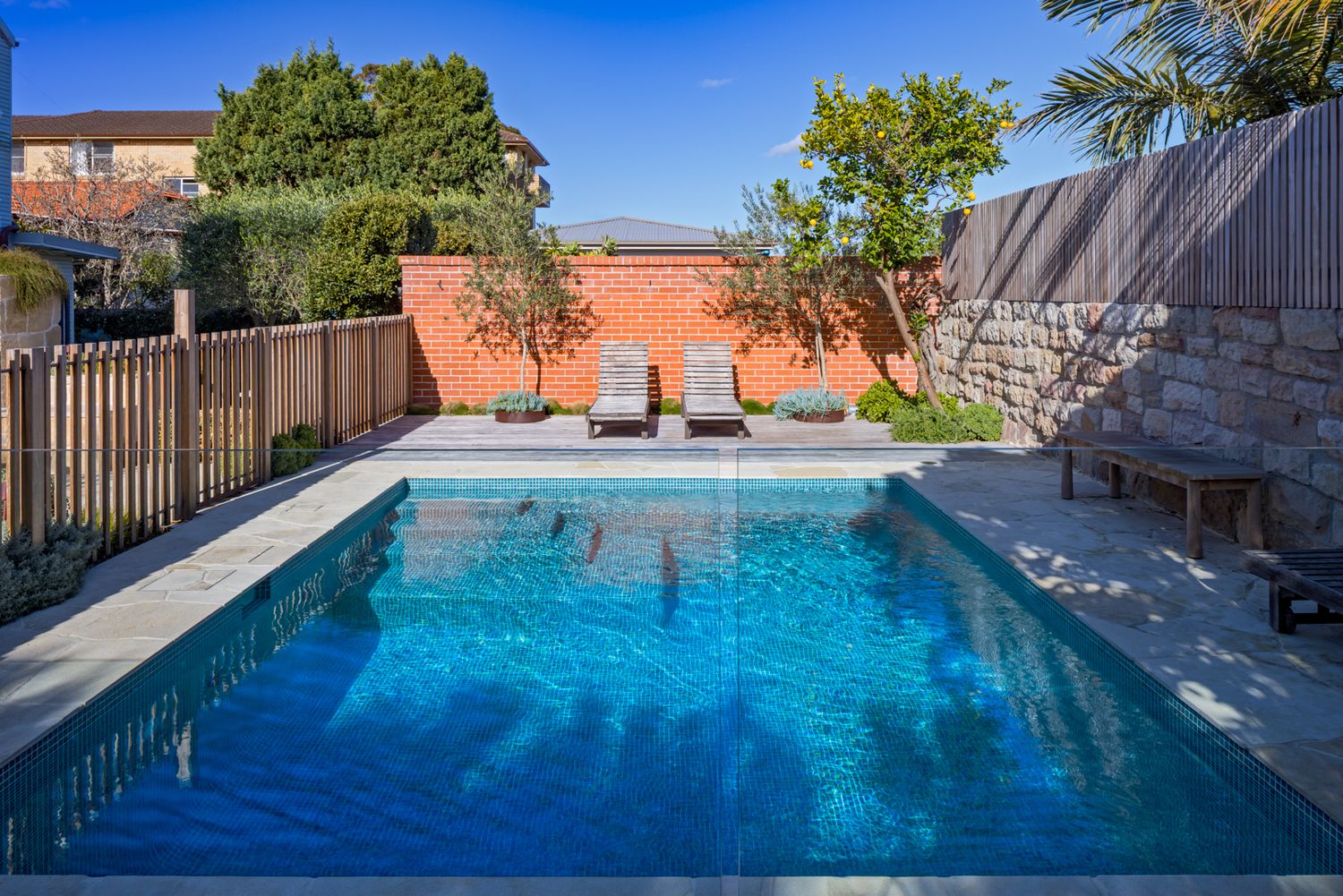 Backyard swimming pool with clear blue water, two lounge chairs, a terracotta wall, timber fence and stone paving.