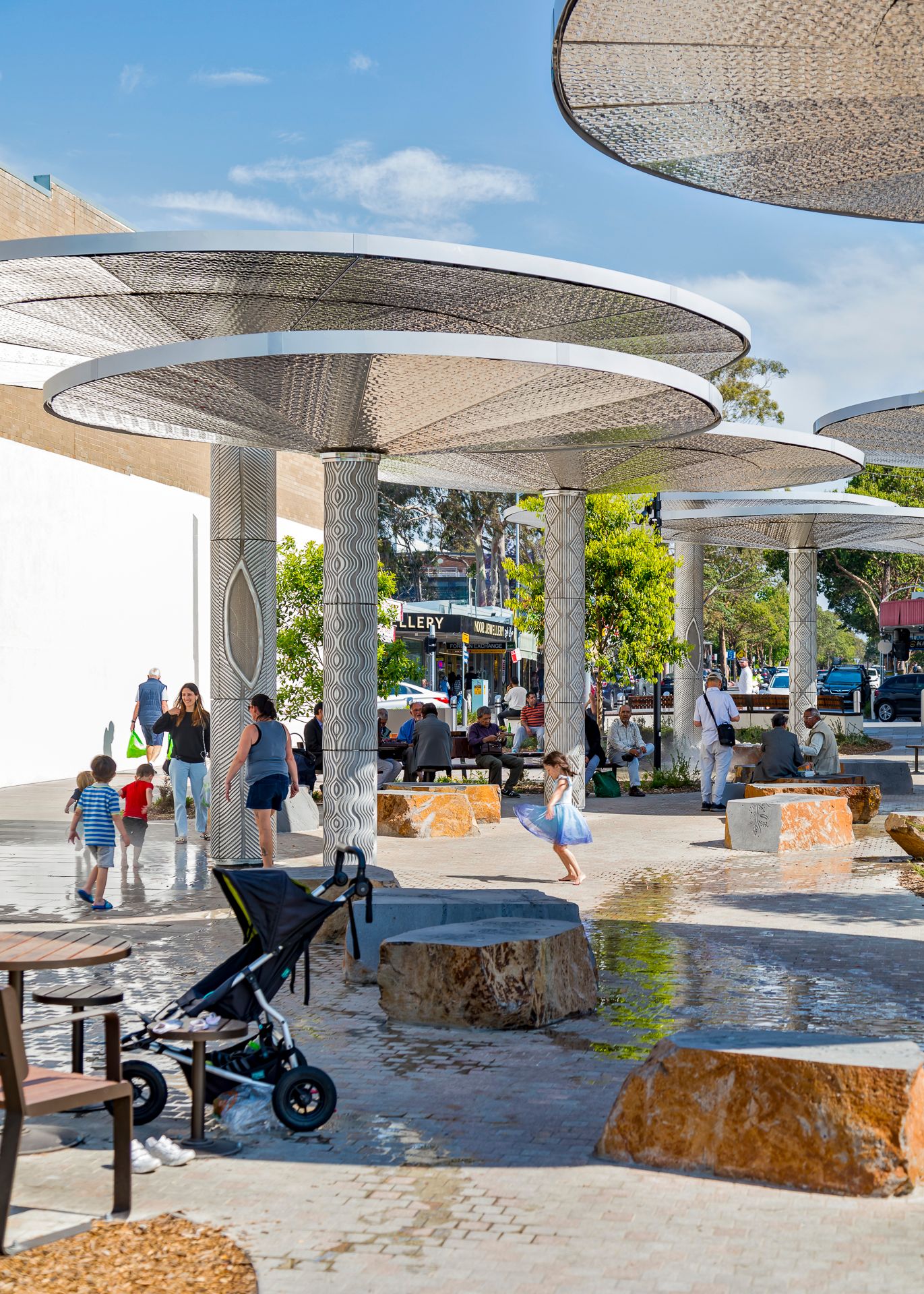 Families and children walk beneath mushroom-shaped shade canopies in a sunny plaza with a shallow water feature and rock seating.