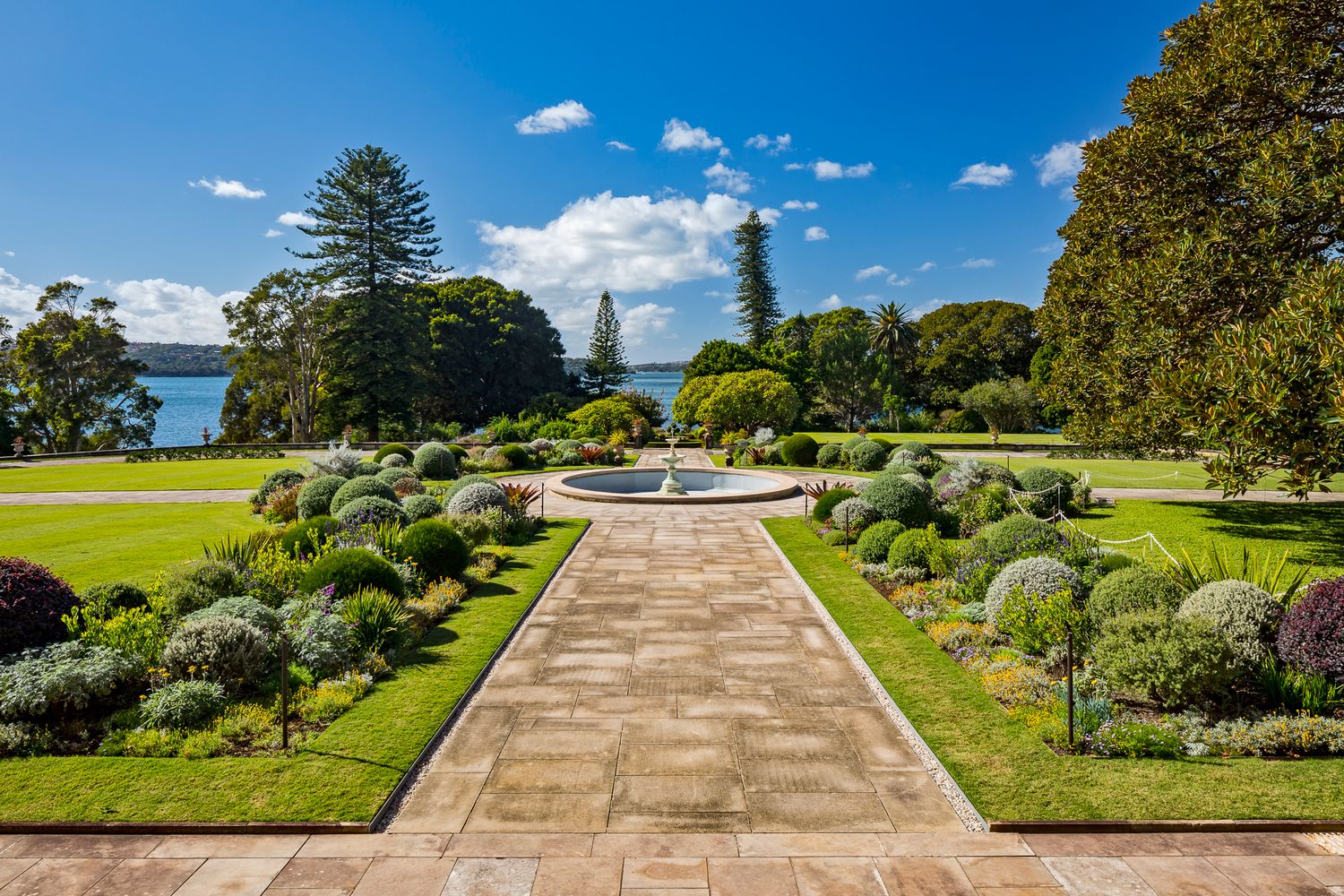 Formal garden with a central fountain, clipped shrubs, and a brick path overlooking a sparkling harbor under blue sky.