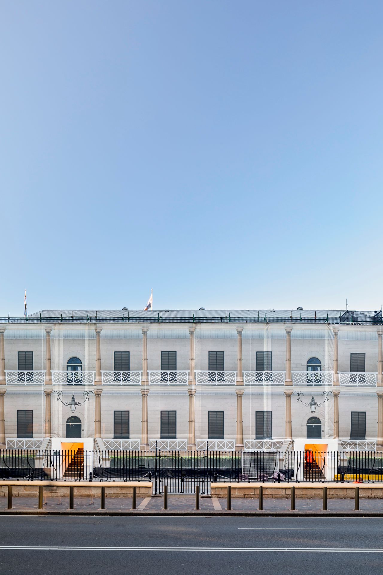 Symmetrical neoclassical building with columns and balconies covered in sheeting, with orange entrance stairs behind a black iron fence.