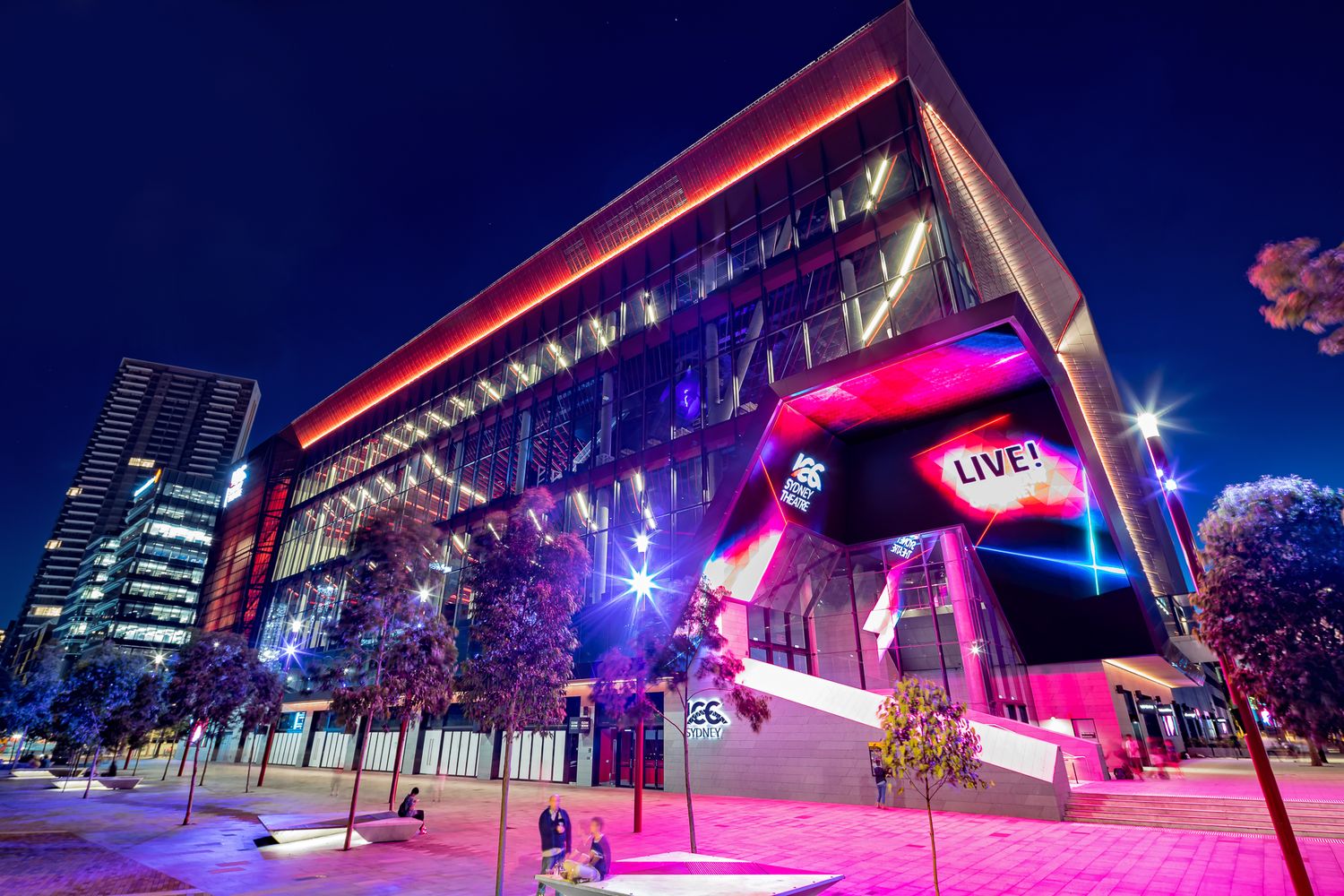 Illuminated glass convention centre at night with colorful pink and purple lighting, Live signage and tree-lined walkways.