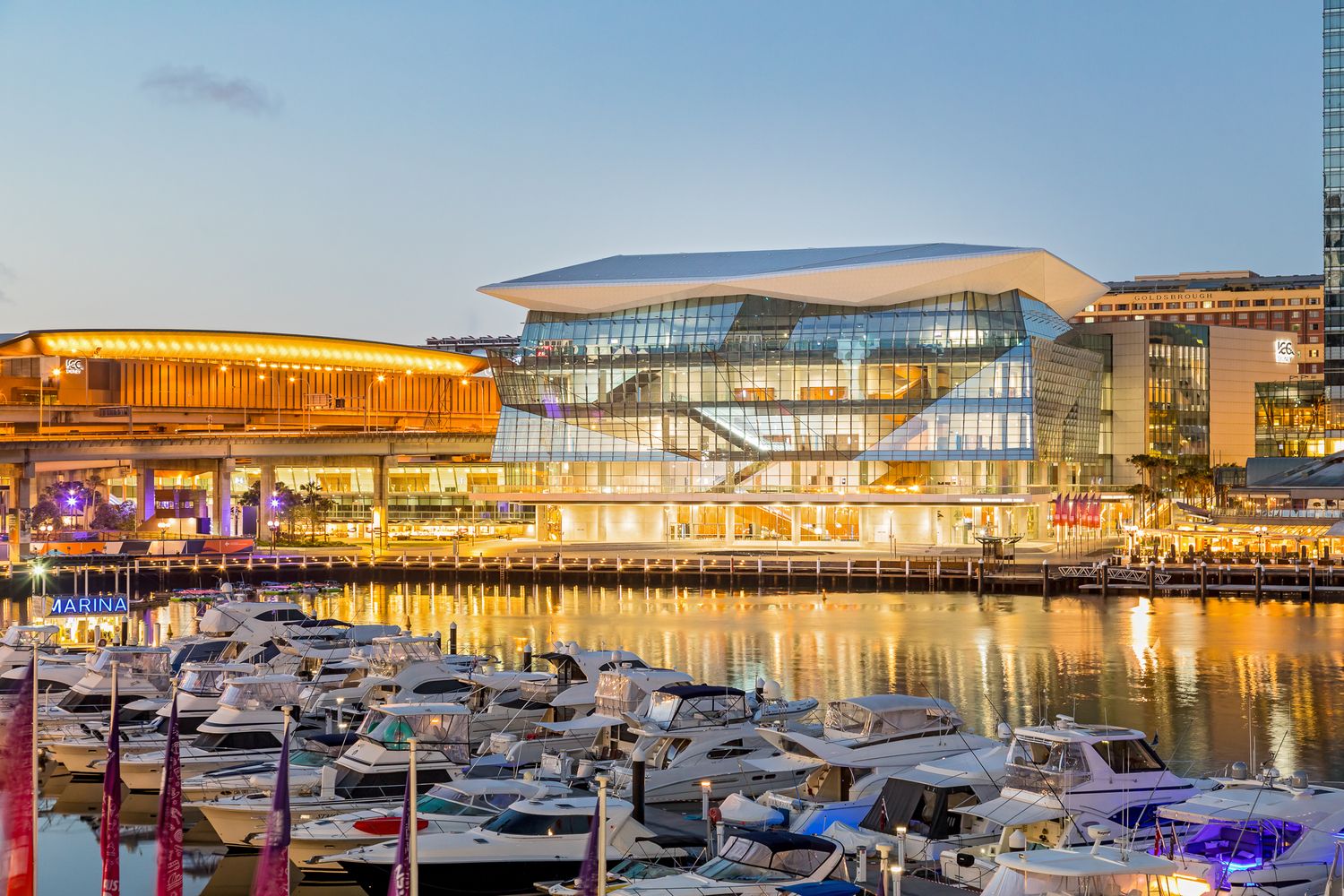 Illuminated waterfront convention centre at dusk with moored yachts and boats lining the marina in the foreground.