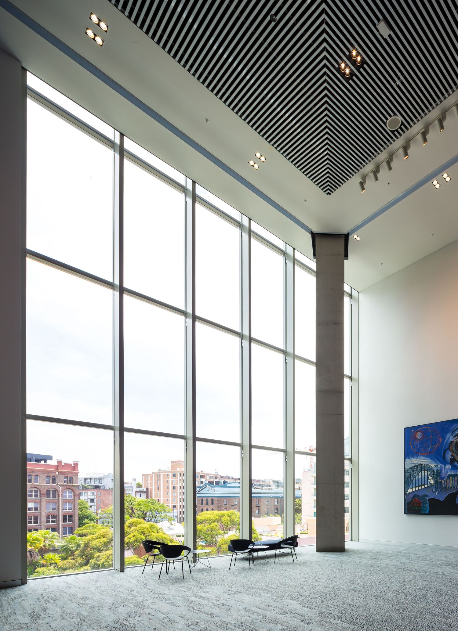 Tall glass curtain wall in a spacious lobby with black chairs, a slatted ceiling, and city views beyond.