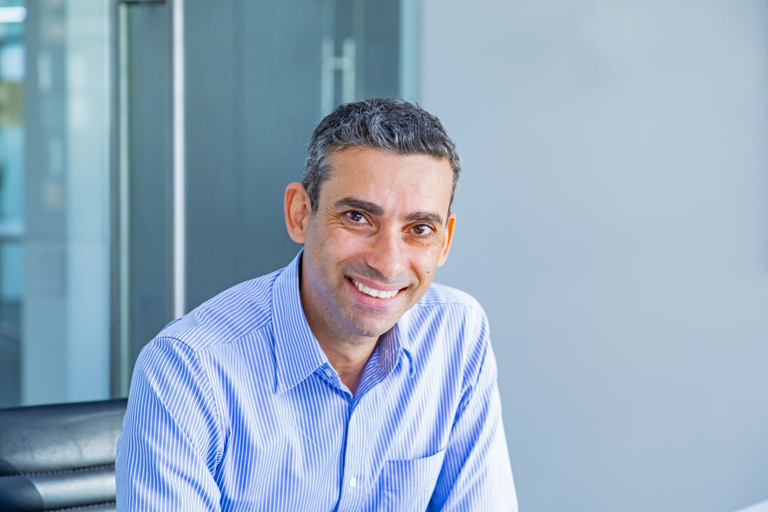 Smiling man with short dark hair in a light blue striped shirt sits in a bright modern office with glass walls.