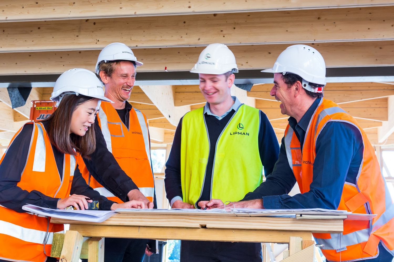 Four construction professionals in hard hats and hi-vis vests review building plans together on a table inside a timber-framed structure.