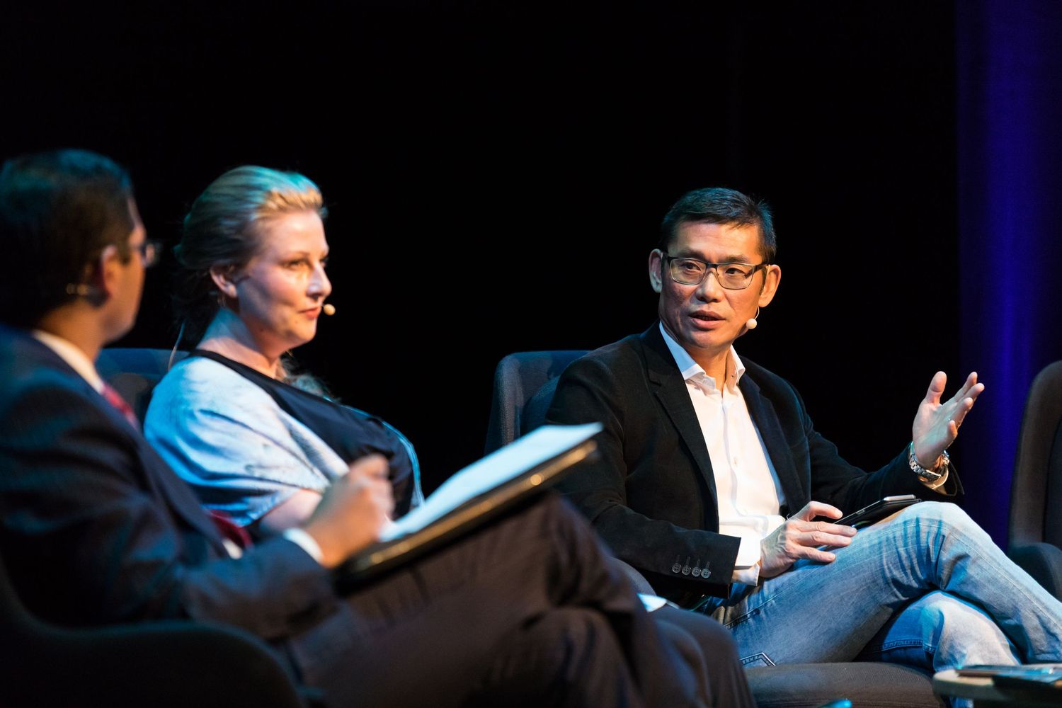 Three people sit on stage in a panel discussion, with a man in glasses gesturing as he speaks into a headset microphone.
