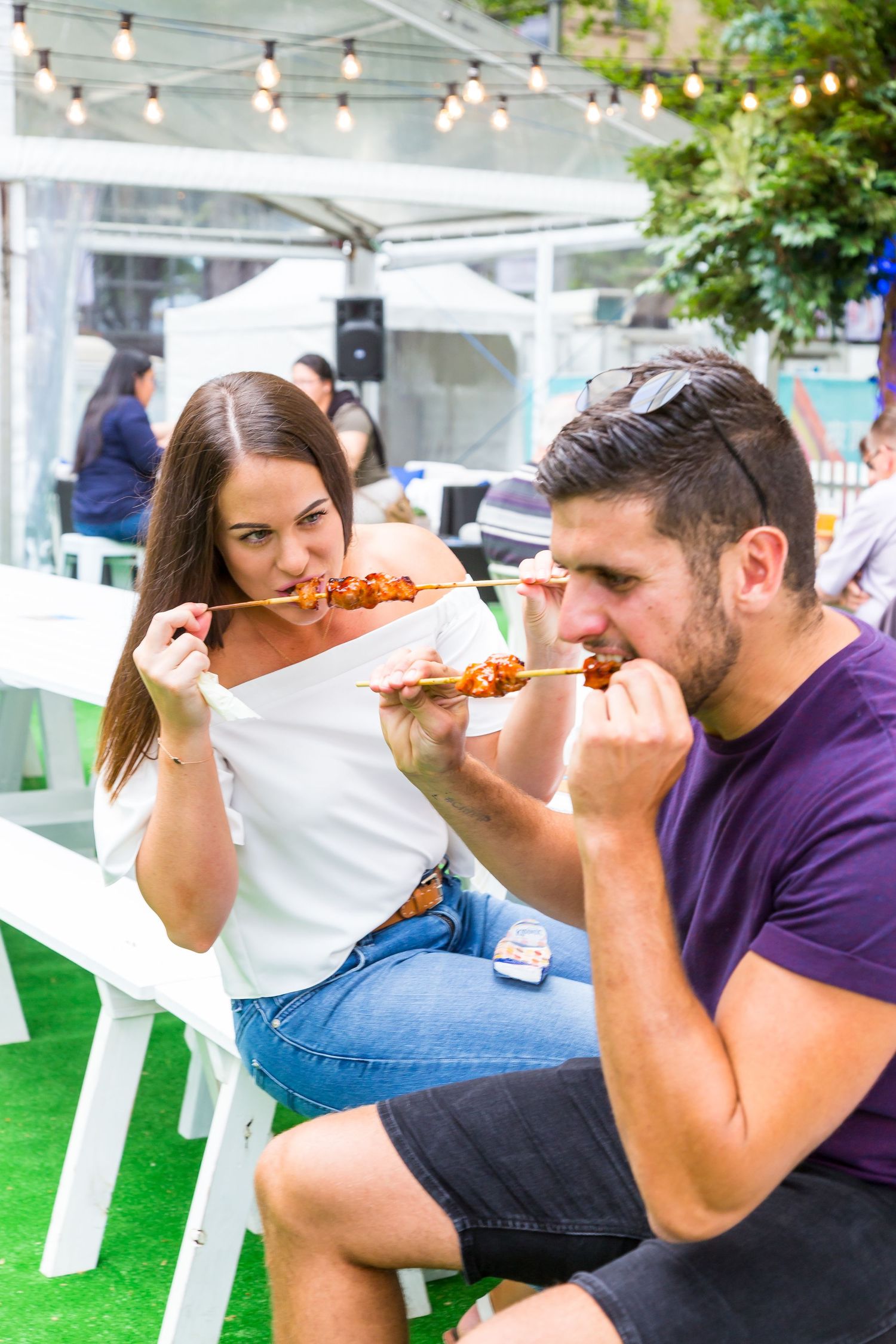 A woman and a man sit on a white bench at an outdoor event, happily eating grilled meat skewers under string lights.