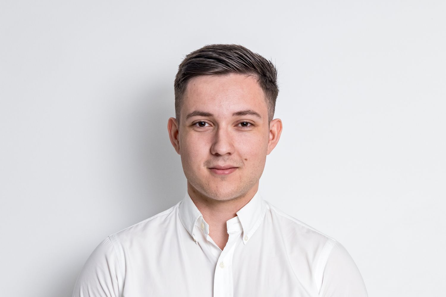Young man with short dark hair wearing a white button-up shirt posing against a plain light background.