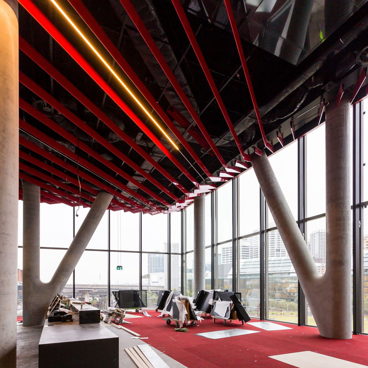 Interior space with angled concrete columns, red ceiling beams and floor-to-ceiling windows overlooking the city.
