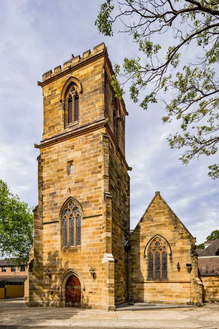 Historic Gothic Revival sandstone church with a square bell tower, pointed arched windows and a red wooden door beneath leafy trees.