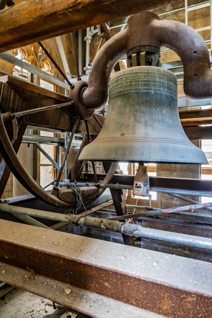 Large vintage bronze church bell mounted in a timber belfry frame with a metal wheel, steel beams and light from arched openings.