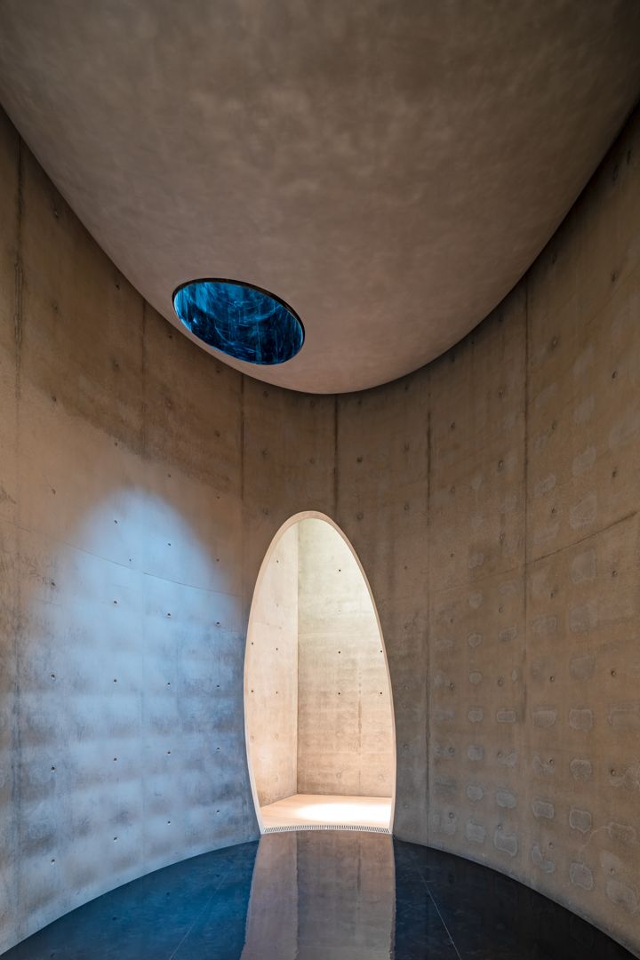 Curved concrete chamber with an arched doorway, circular skylight opening to the sky and a reflective dark floor.
