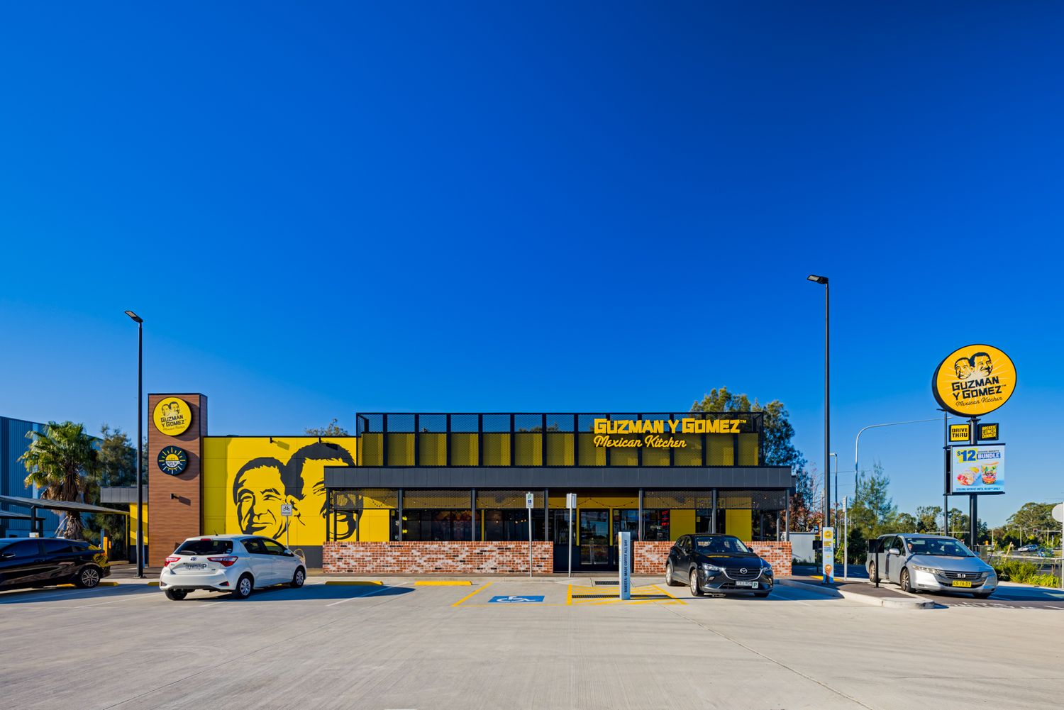 Guzman y Gomez Mexican Kitchen drive-through restaurant with yellow facade, mural, parked cars and a bright blue sky.