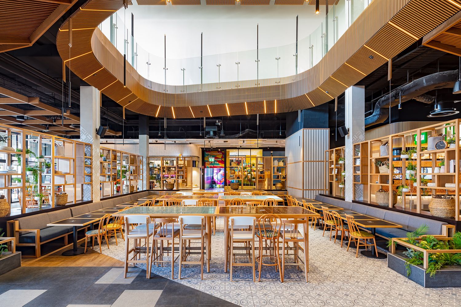 Spacious modern food hall with wooden tables, bar stools, timber shelving with plants and a curved wooden ceiling feature.