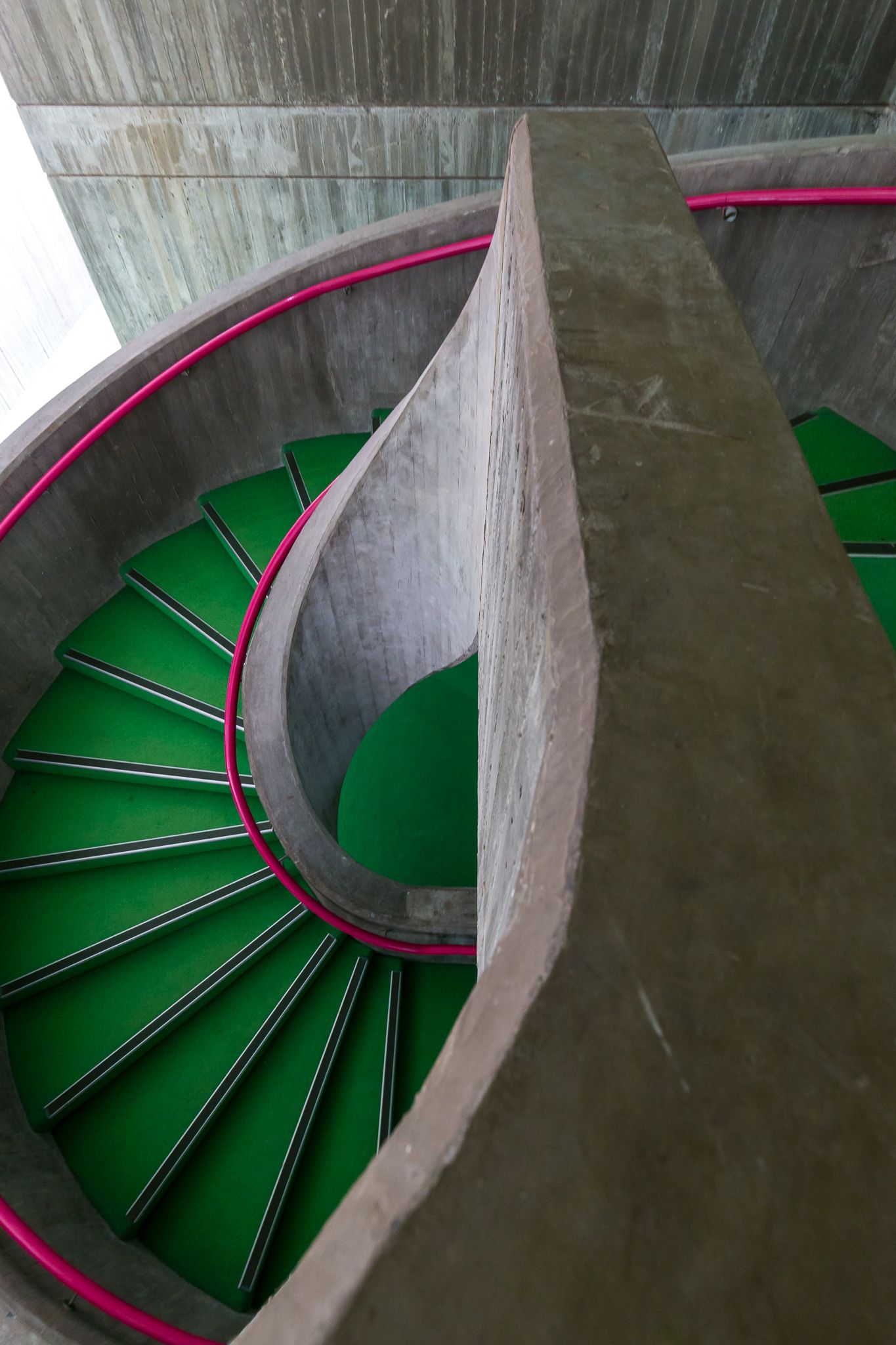 Overhead view of a curved concrete spiral staircase with green treads and bright pink handrails.