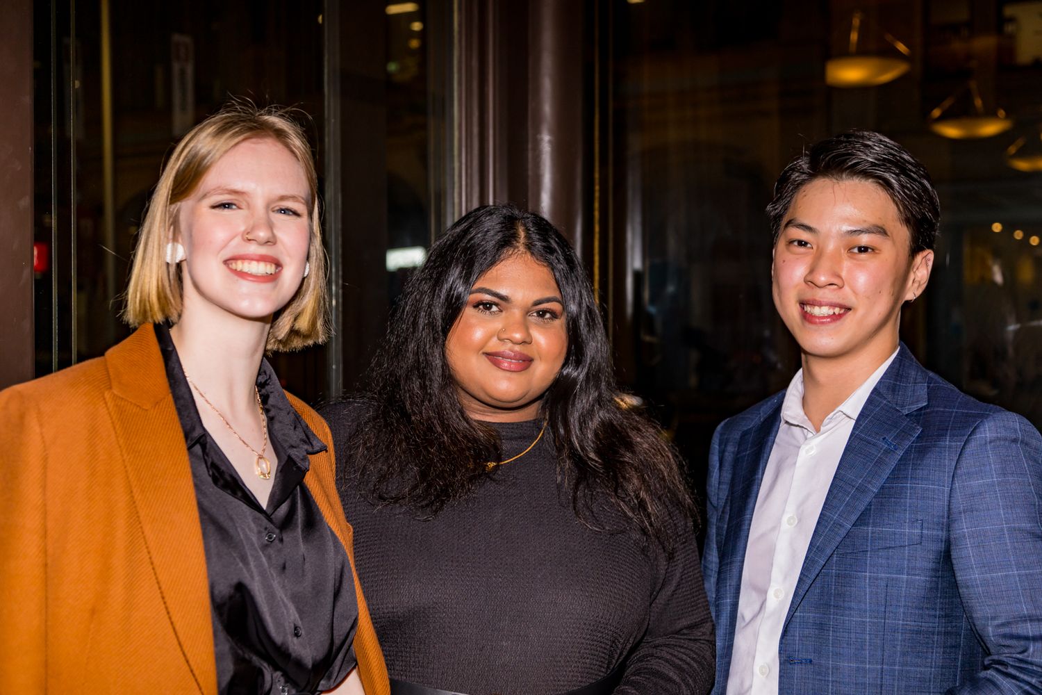 Three smiling colleagues pose together at an indoor evening event, wearing an orange blazer, a dark top and a blue suit jacket.