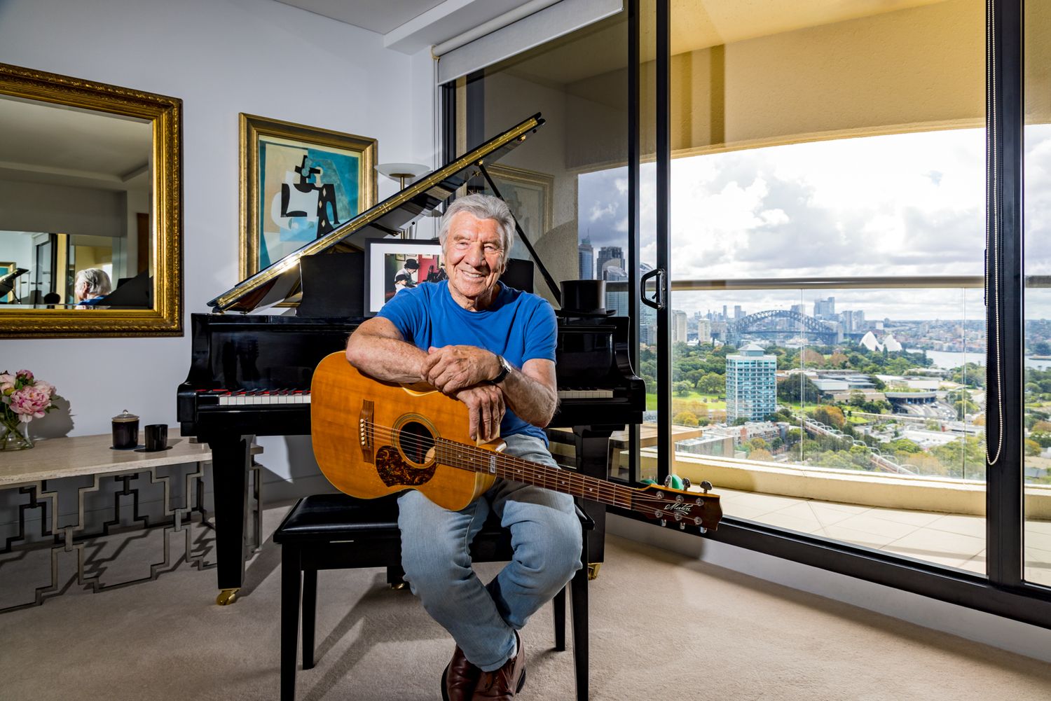 A smiling man in a blue shirt and jeans sits holding an acoustic guitar beside a black grand piano in an apartment with city views.
