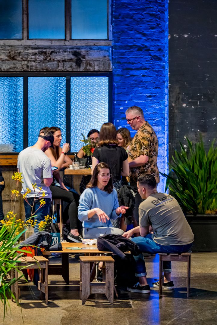 Group of people chat around wooden tables in a casual venue with blue-lit brick walls, large windows and green plants.