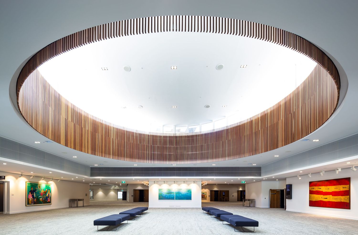 Airy foyer with a large oval timber-lined skylight, blue benches and colourful artworks on the walls of a modern venue.