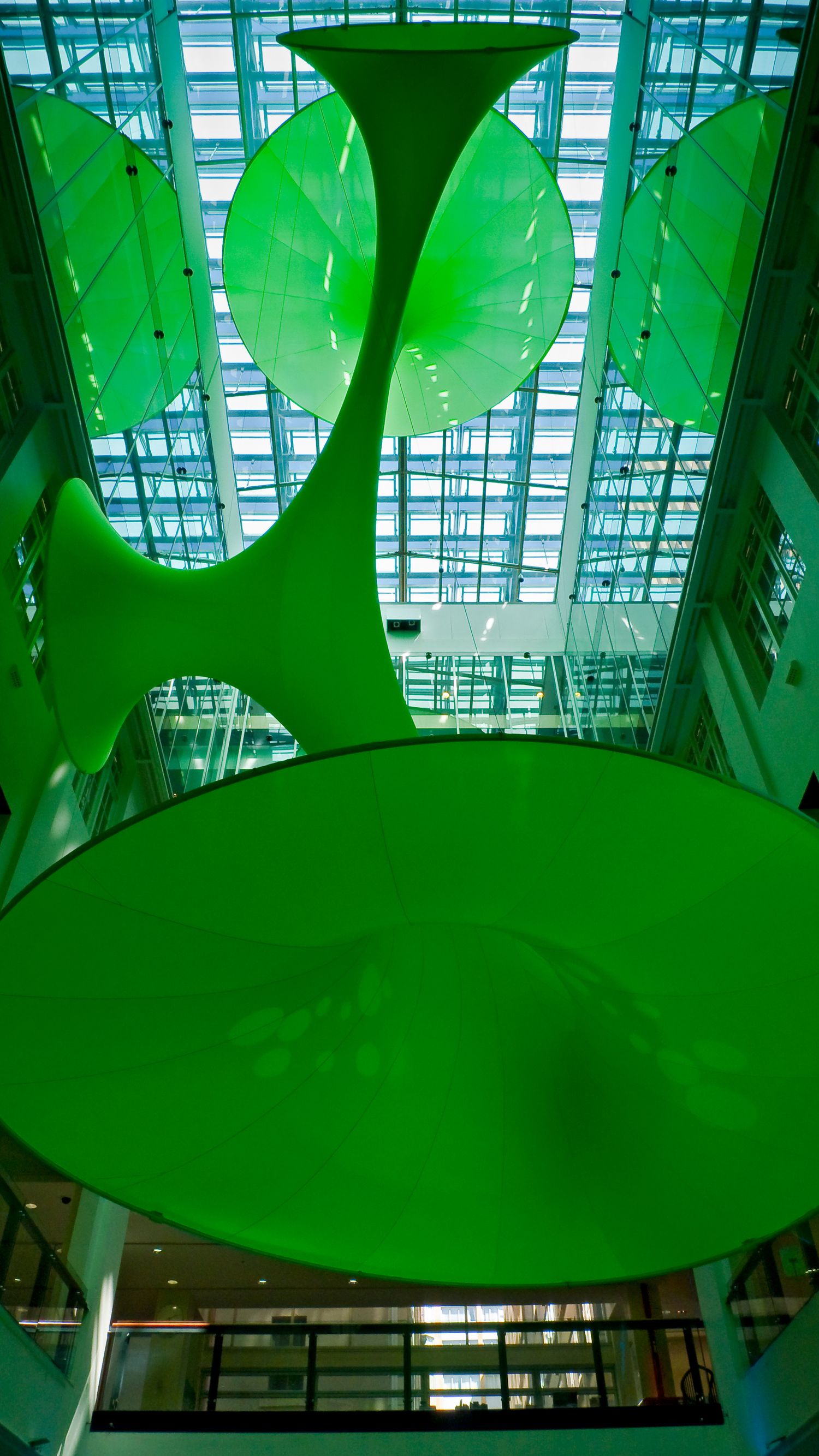 Green suspended fabric sculptures with trumpet-shaped forms hang in a glass-roofed atrium with a steel-framed skylight above.