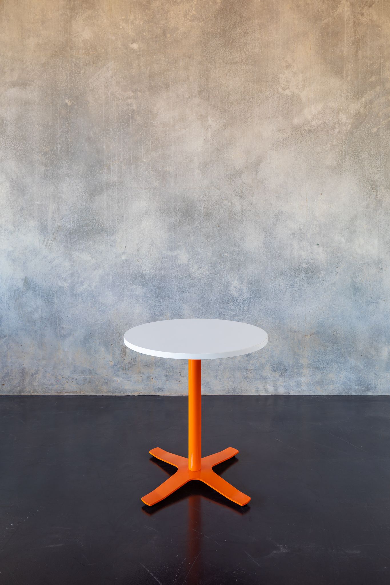 Small round café table with a white top and orange cross-shaped metal base on a dark floor against a concrete wall.