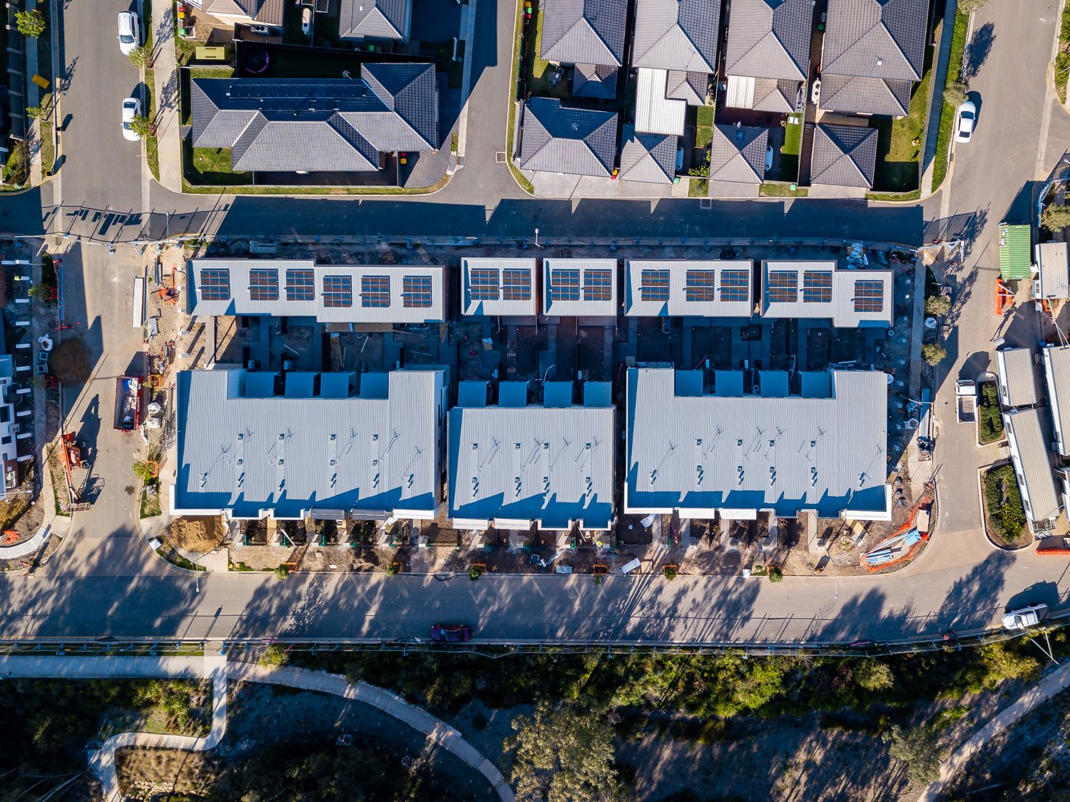 Top-down aerial view of a new townhouse row with fresh metal roofing and solar panels beside a street and bushland.