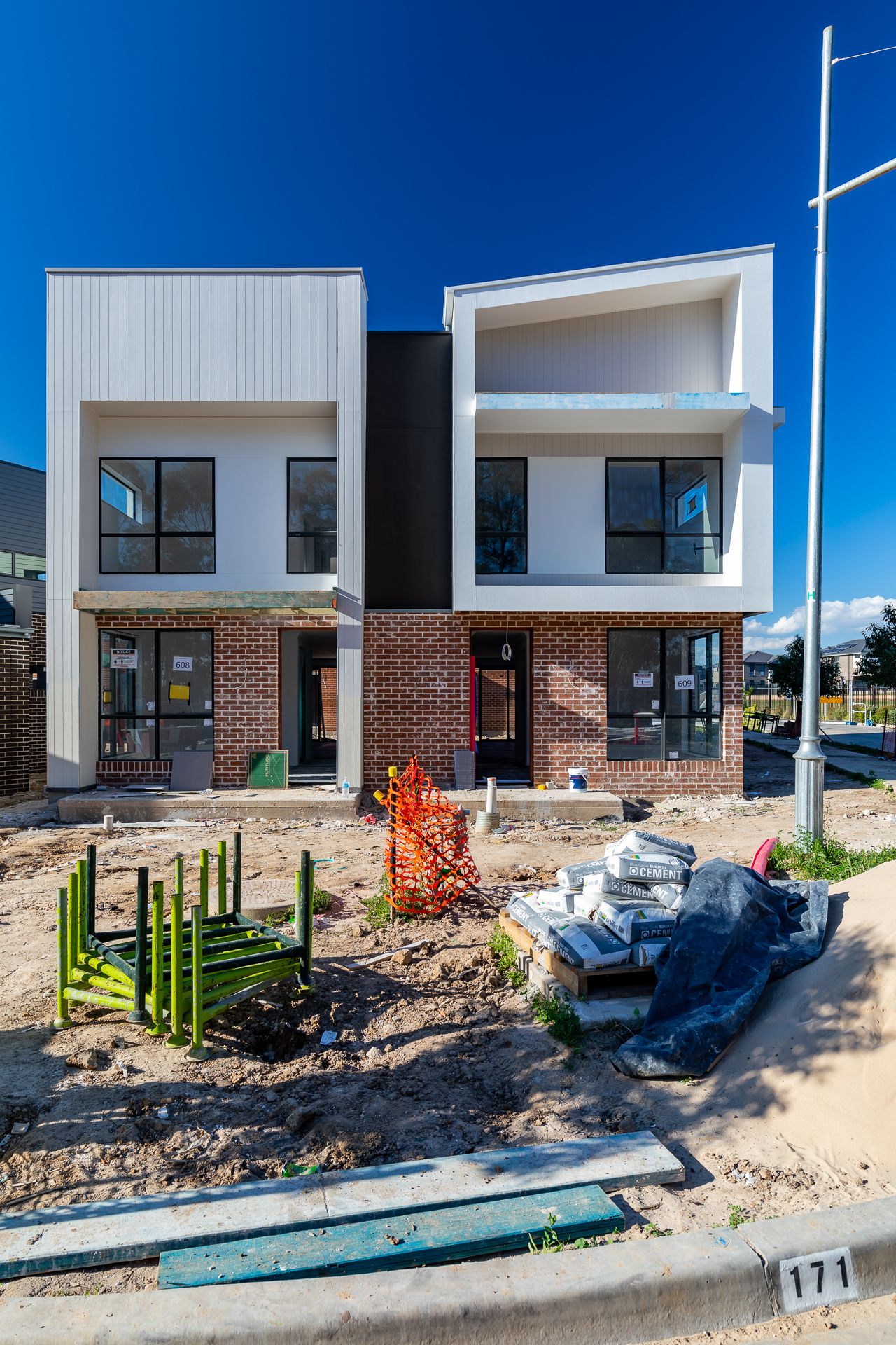 Two-storey modern duplex homes with white render and brick facades nearing completion, with building materials stacked out front on site.