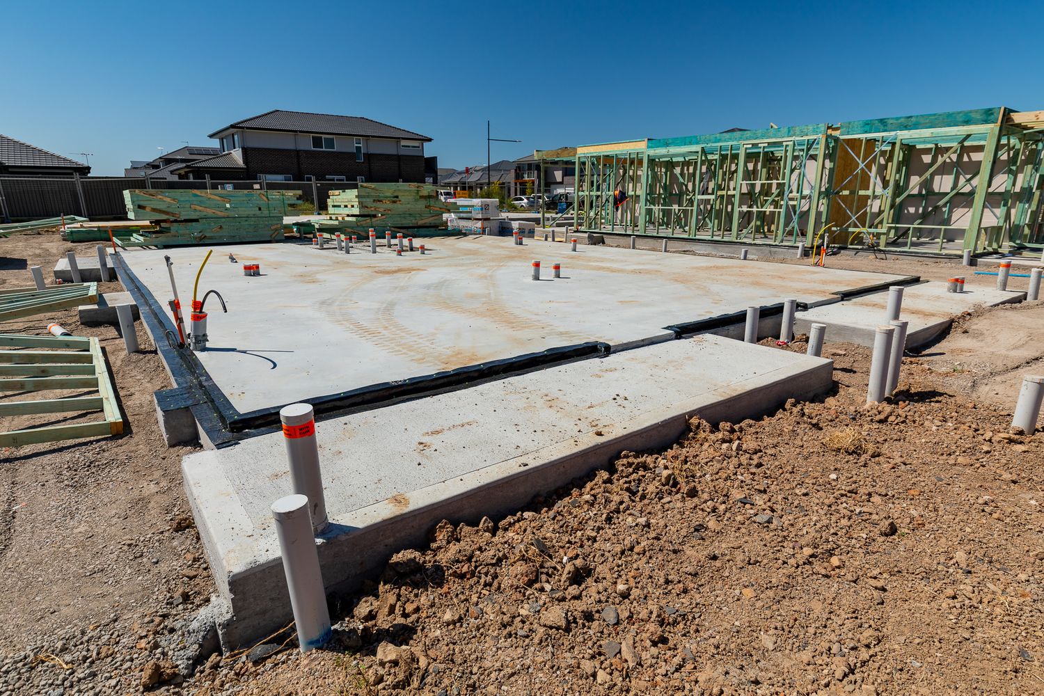 Concrete slab foundations poured at a residential construction site with timber house framing and neighboring homes under a clear blue sky.