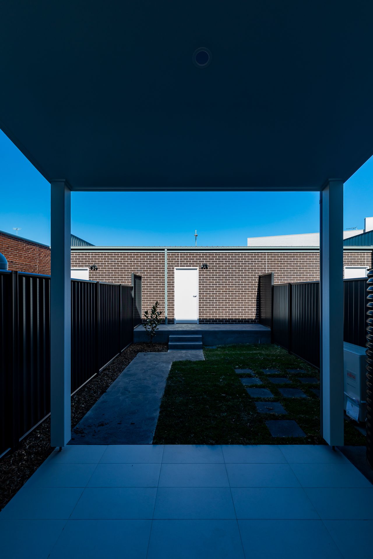 View from a covered patio across a landscaped backyard with new turf, stepping stones, black fencing and a brick rear building.