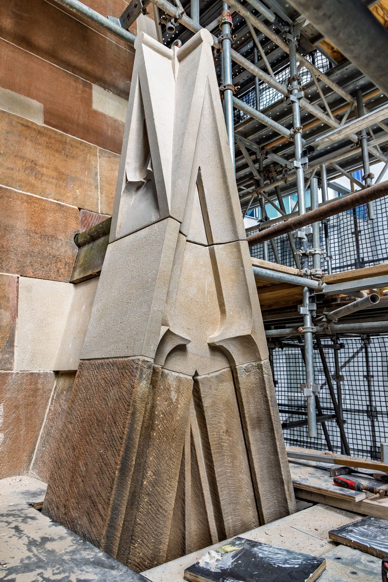 Carved sandstone pinnacle with geometric Gothic detailing standing beside restored stone walls under construction scaffolding.