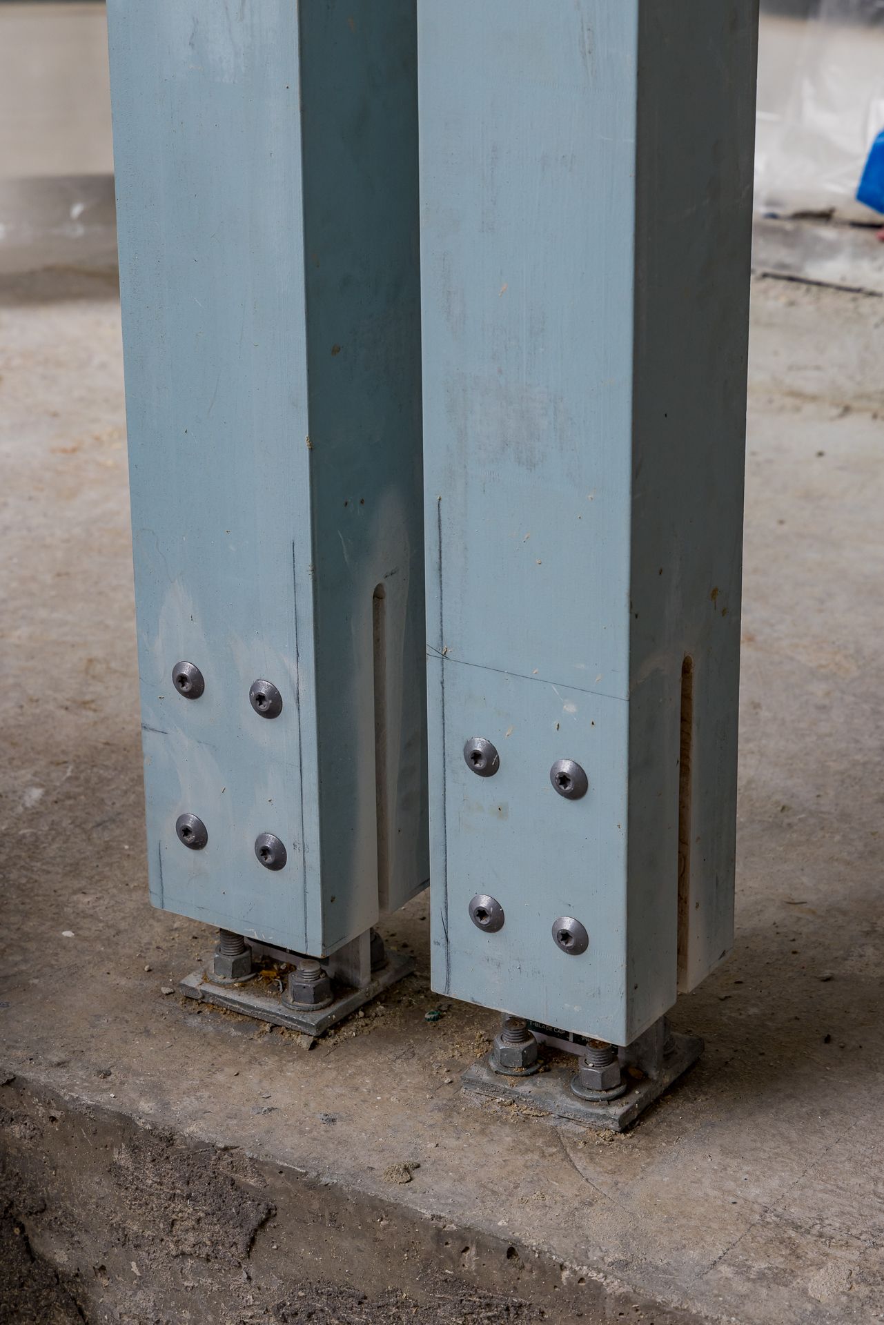 Close-up of two blue-gray steel columns bolted to a concrete slab with base plates at a building construction site.