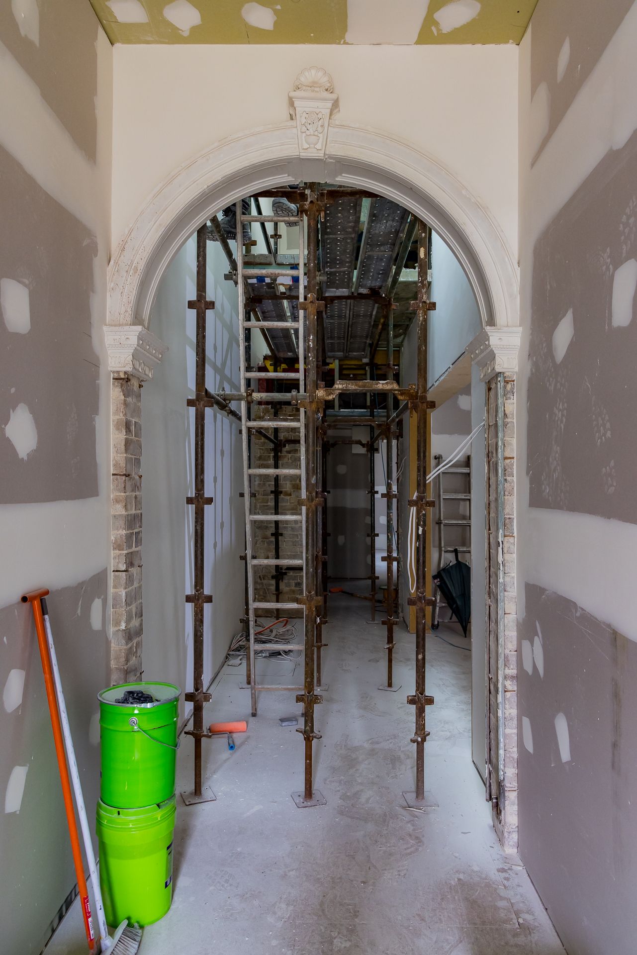 Arched hallway with ornate plaster moulding, exposed brick pillars and scaffolding inside a home under renovation.