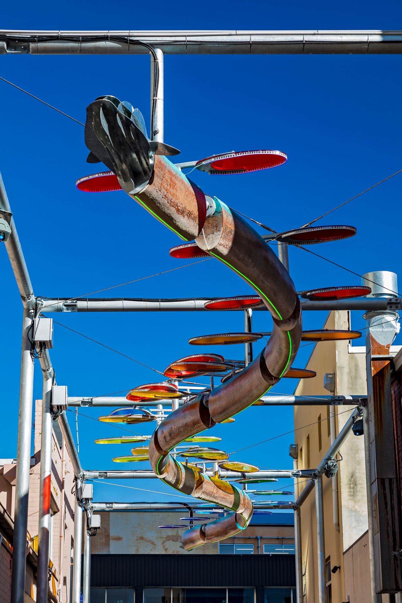 Long snaking bronze fish sculpture with red, yellow and green discs hangs from steel frames above a laneway with brick buildings.