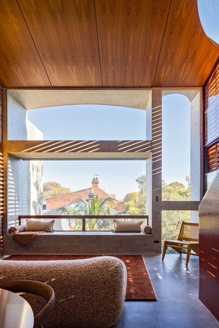 Built-in window seat with cushions in a timber-ceilinged room, framing sunlit views of rooftops, palms and trees.