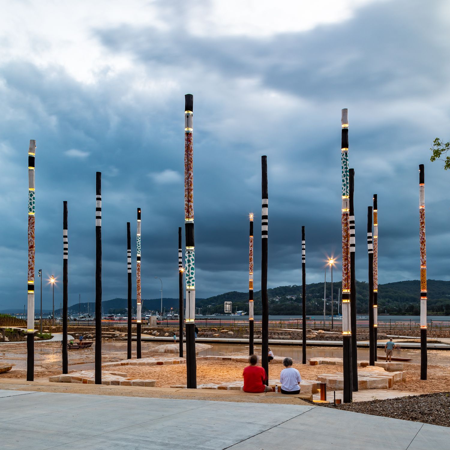 Visitors sit on a sandy plaza surrounded by tall patterned art poles overlooking a harbour under dramatic stormy clouds.